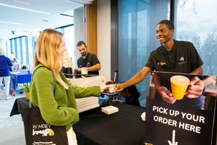 Person handing a takeaway coffee to a customer at a pickup counter with a sign reading ‘Pick up your order here.
