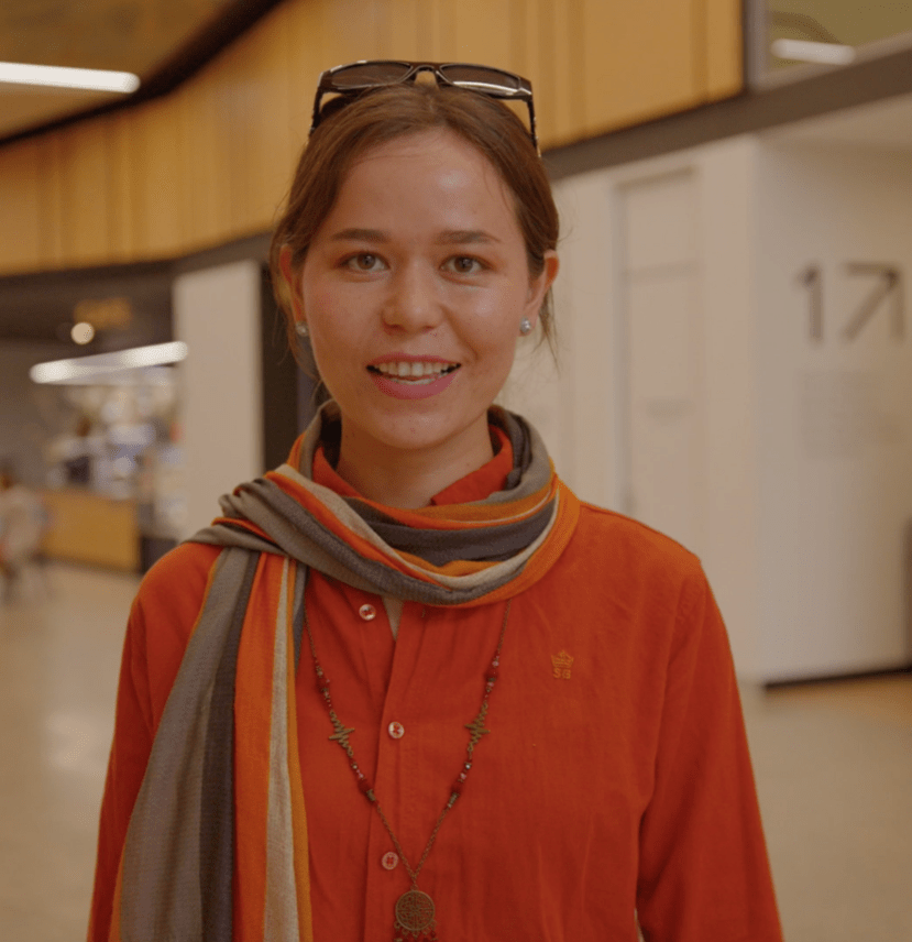 Nazdana wearing an orange shirt with a multicoloured scarf and necklace, standing indoors in a modern building.